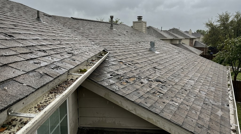 suburban roof with fresh hail impact damage, scattered granules in the gutter, damp surfaces under a heavy grey post-storm sky